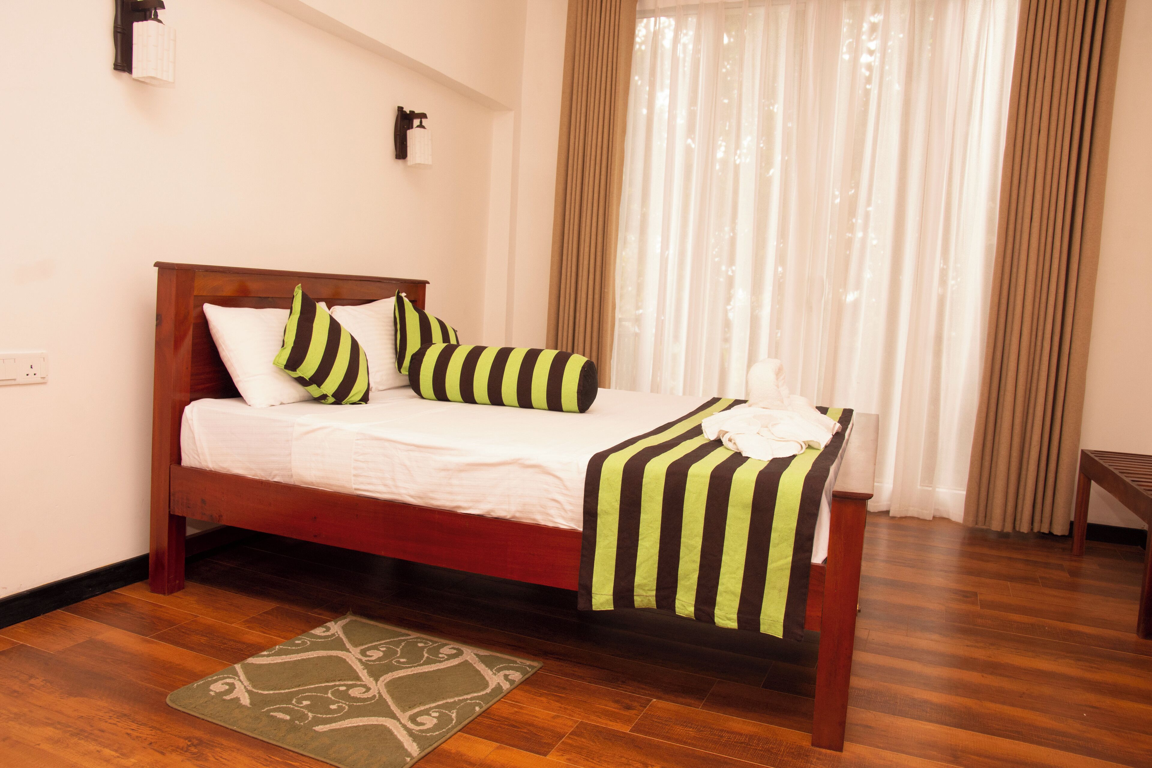 A neatly made bed with striped bedding, a wooden headboard, and a green and white patterned rug on a wooden floor.