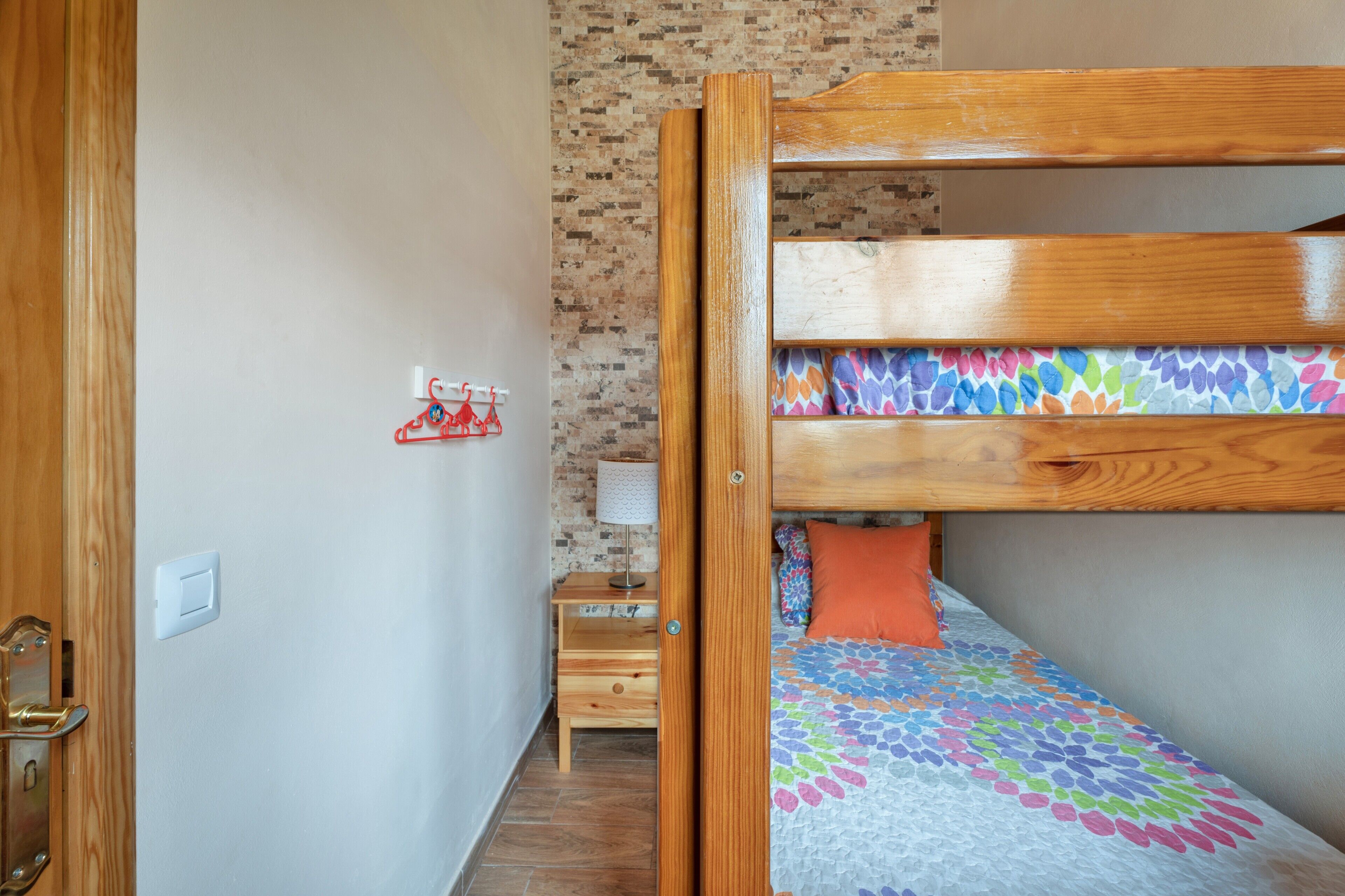 A bunk bed with a colorful quilt, a small wooden nightstand with a lamp, and a wall-mounted red hanger.