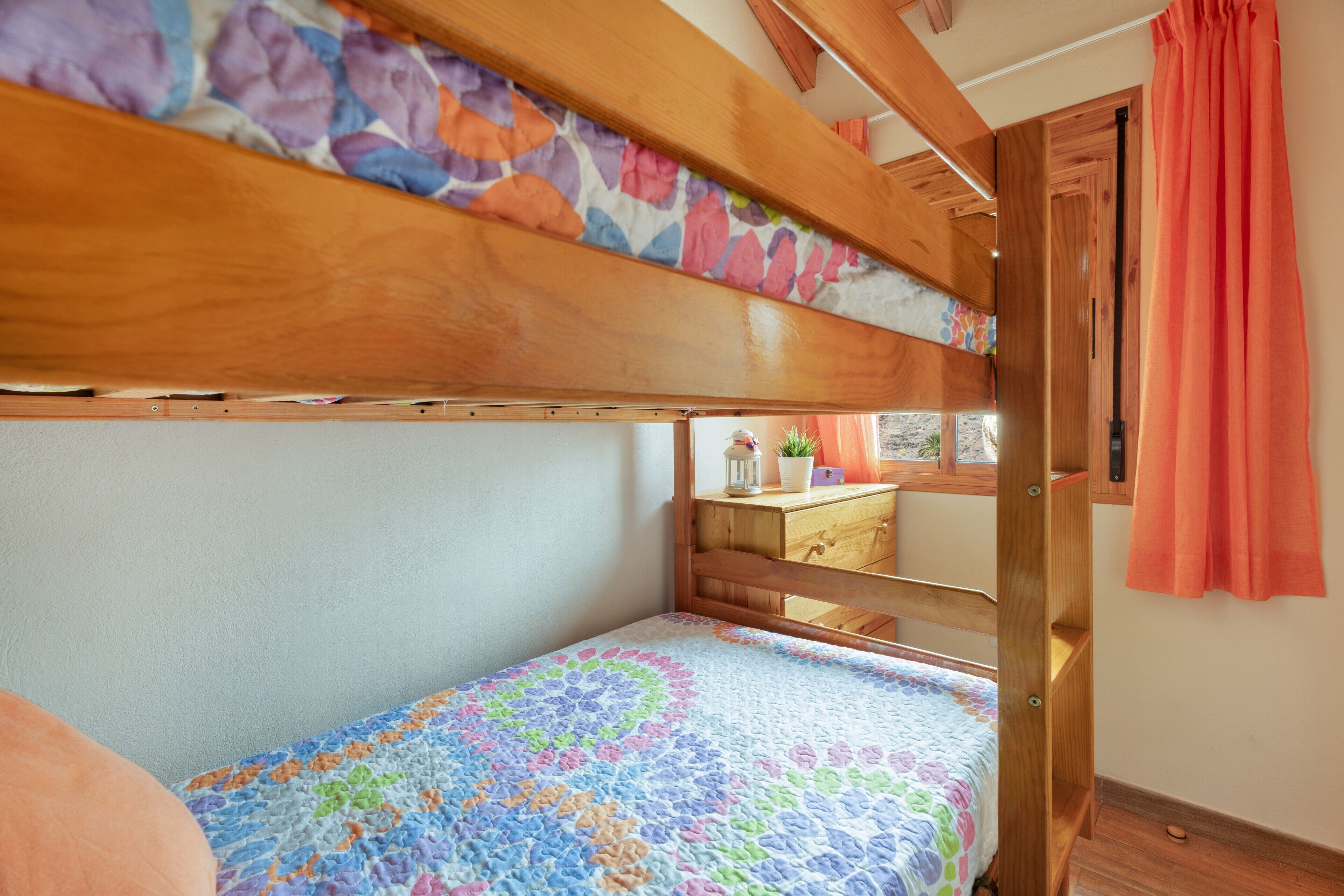 Een stapelbed met een kleurrijke quilt, een houten hoofdbord en een raam met gordijnen.