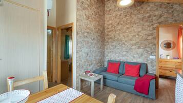 A room with a stone wall, a grey sofa with red cushions, a wooden dining table, and a small white table with a bowl.