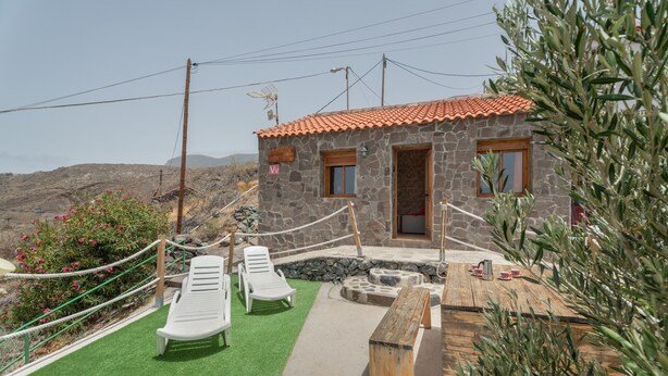 A stone building with a red-tiled roof, a small porch, and a green lawn area with lounge chairs.