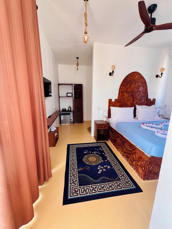 Una habitación de hotel con cama, ventilador de techo, televisor, cabecero de madera, mesita de noche y alfombra decorativa.