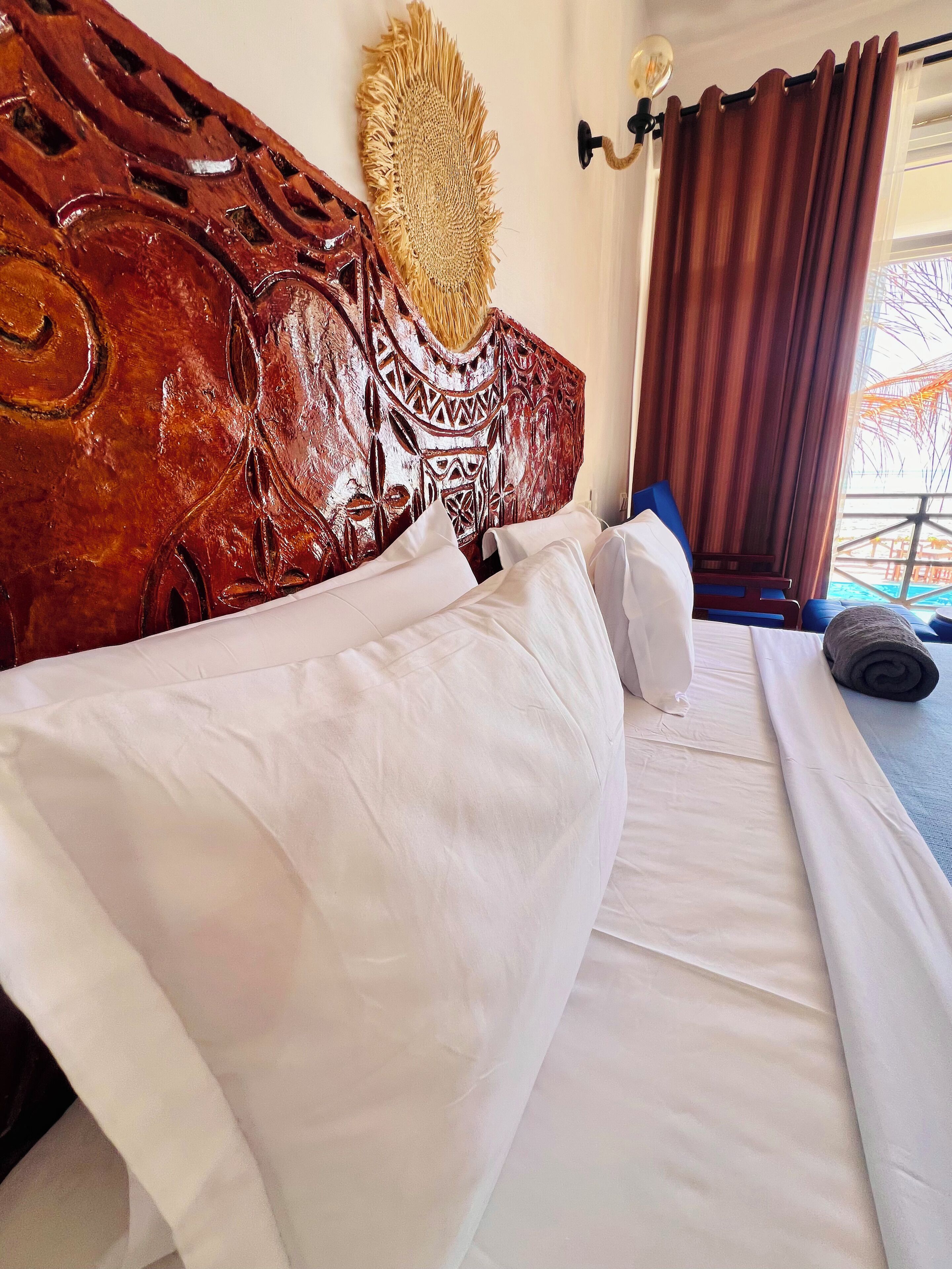 A neatly made bed with white linens, a wooden headboard with intricate carvings, a wall-mounted decorative piece, and a window with curtains.