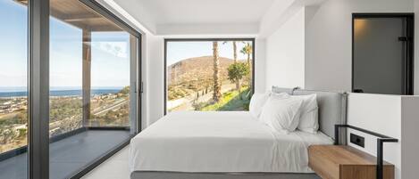 A bedroom with a large bed, a wooden bedside table, and a view of a mountainous landscape through floor-to-ceiling windows.