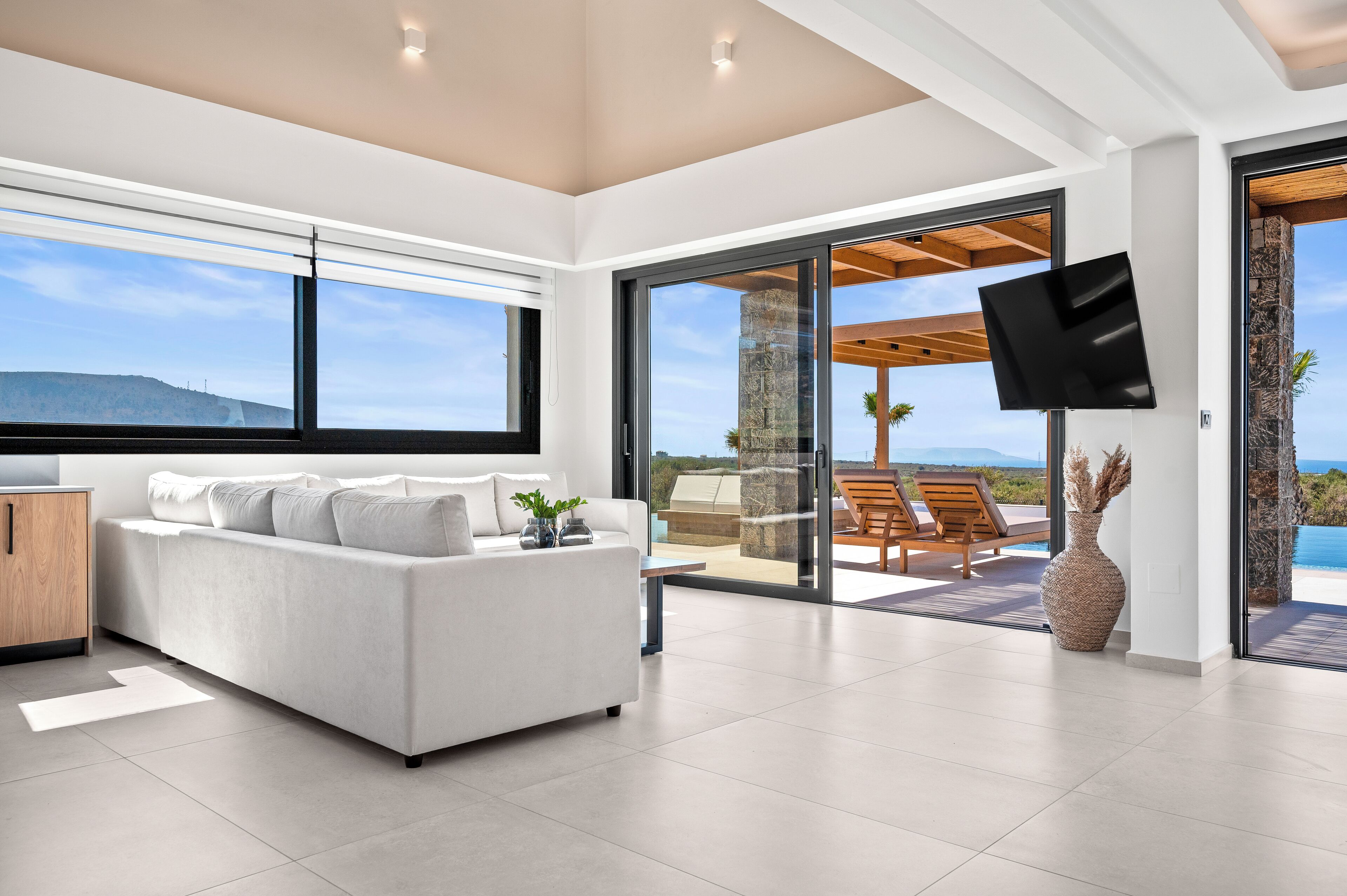 A modern living room with a white sofa, a flat-screen TV mounted on the wall, and a large sliding glass door leading to an outdoor patio with a view of the ocean.