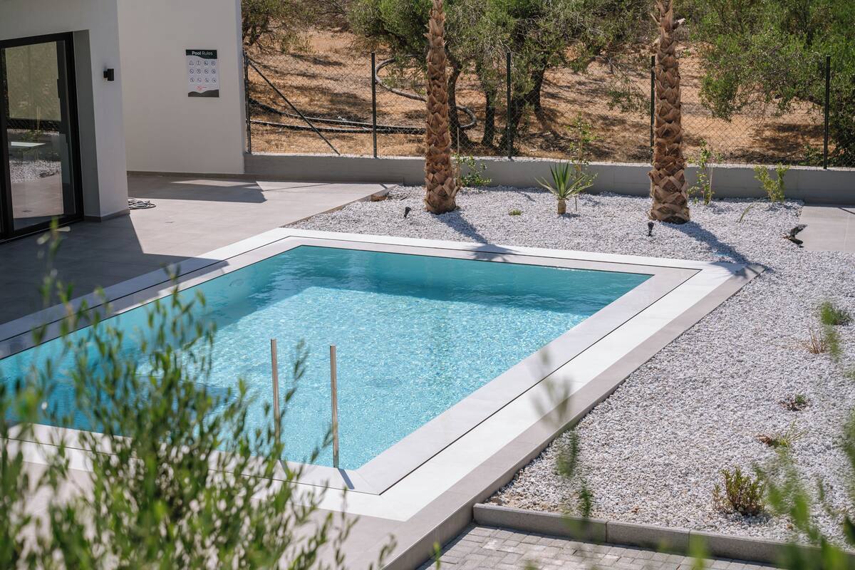 Una piscina rectangular con superficie de agua azul cristalina, rodeada por una zona de grava y palmeras.