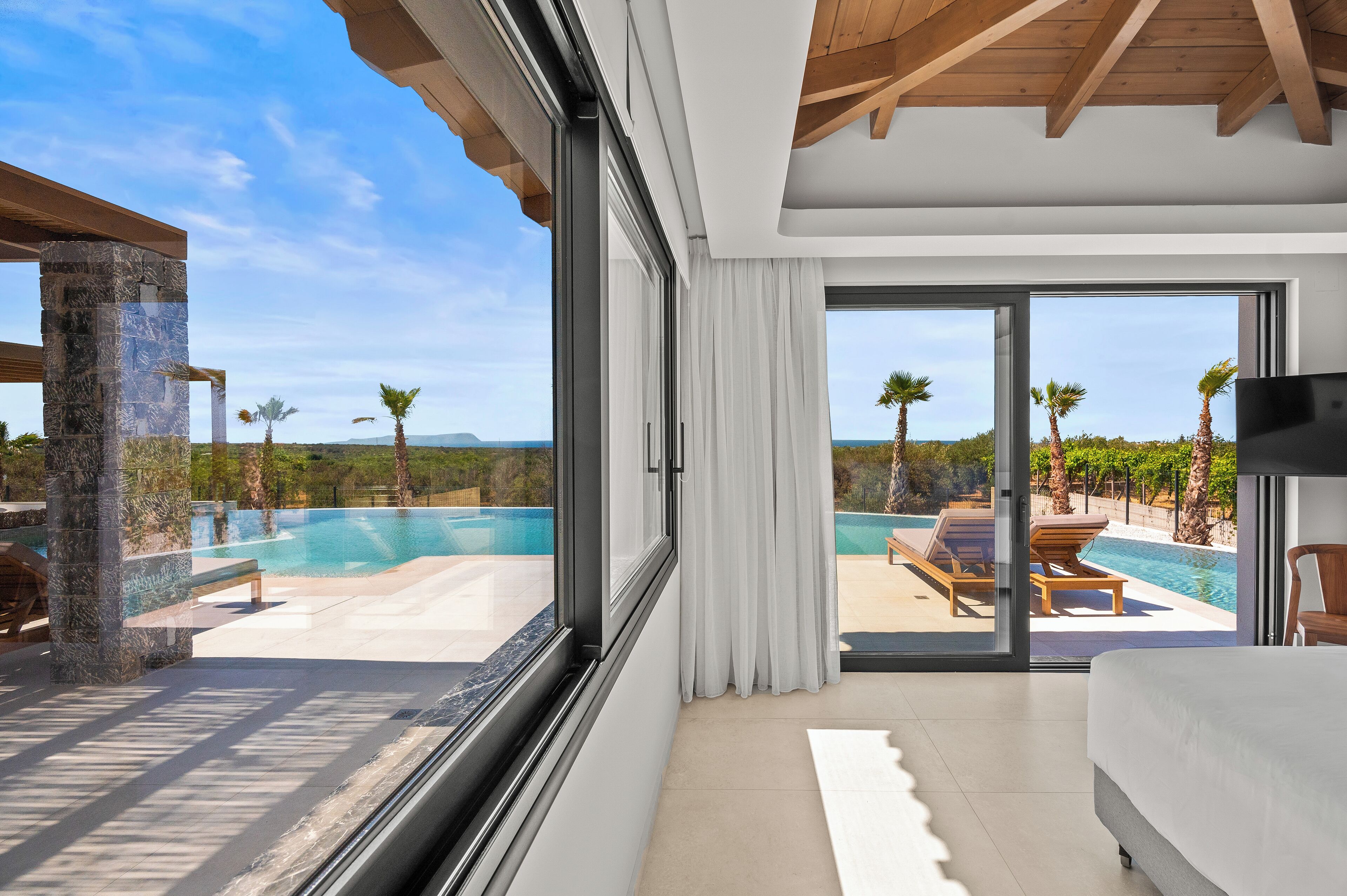 A modern hotel room with a large window, a bed, and a view of a pool and palm trees.