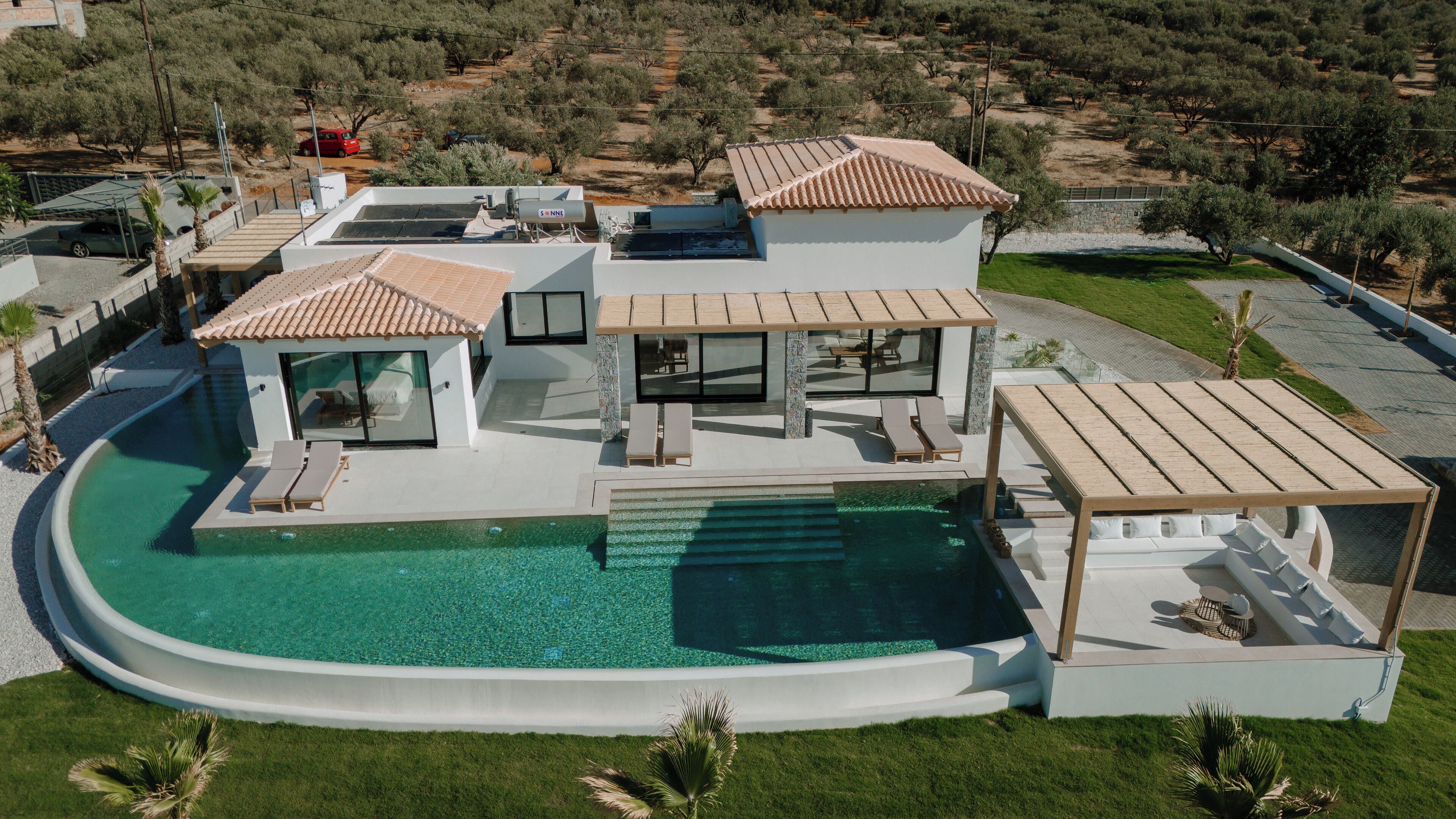 A modern villa with a large swimming pool, multiple outdoor seating areas, and a view of the surrounding landscape.