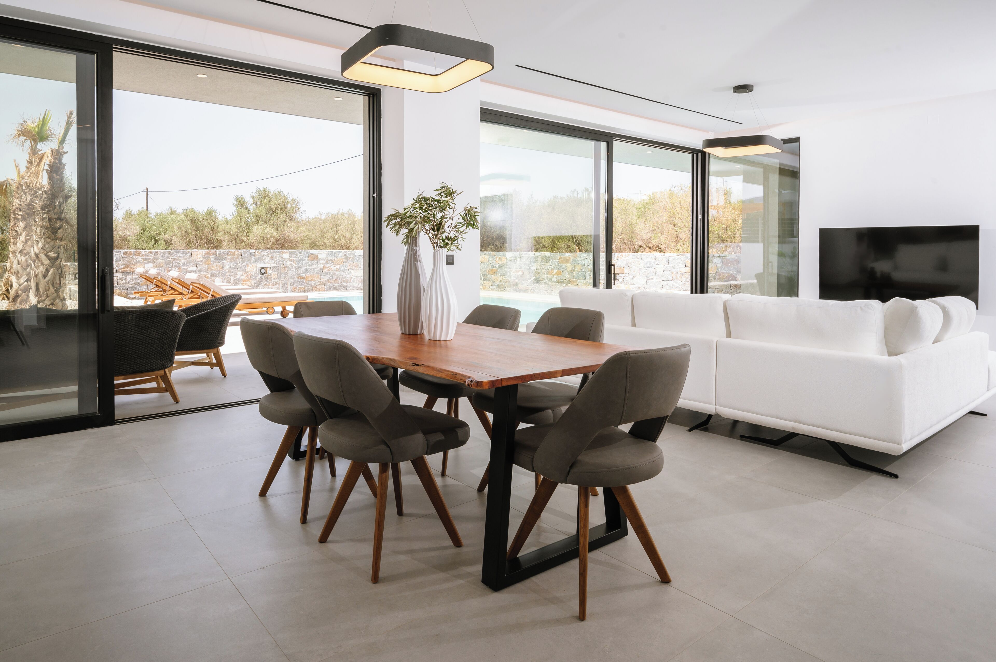 A modern living room with a wooden dining table, grey chairs, a white sofa, and a TV.