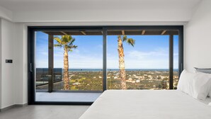A bedroom with a large bed, a view of palm trees, and a cityscape through floor-to-ceiling windows. - Temma Villas (Hersonissos)