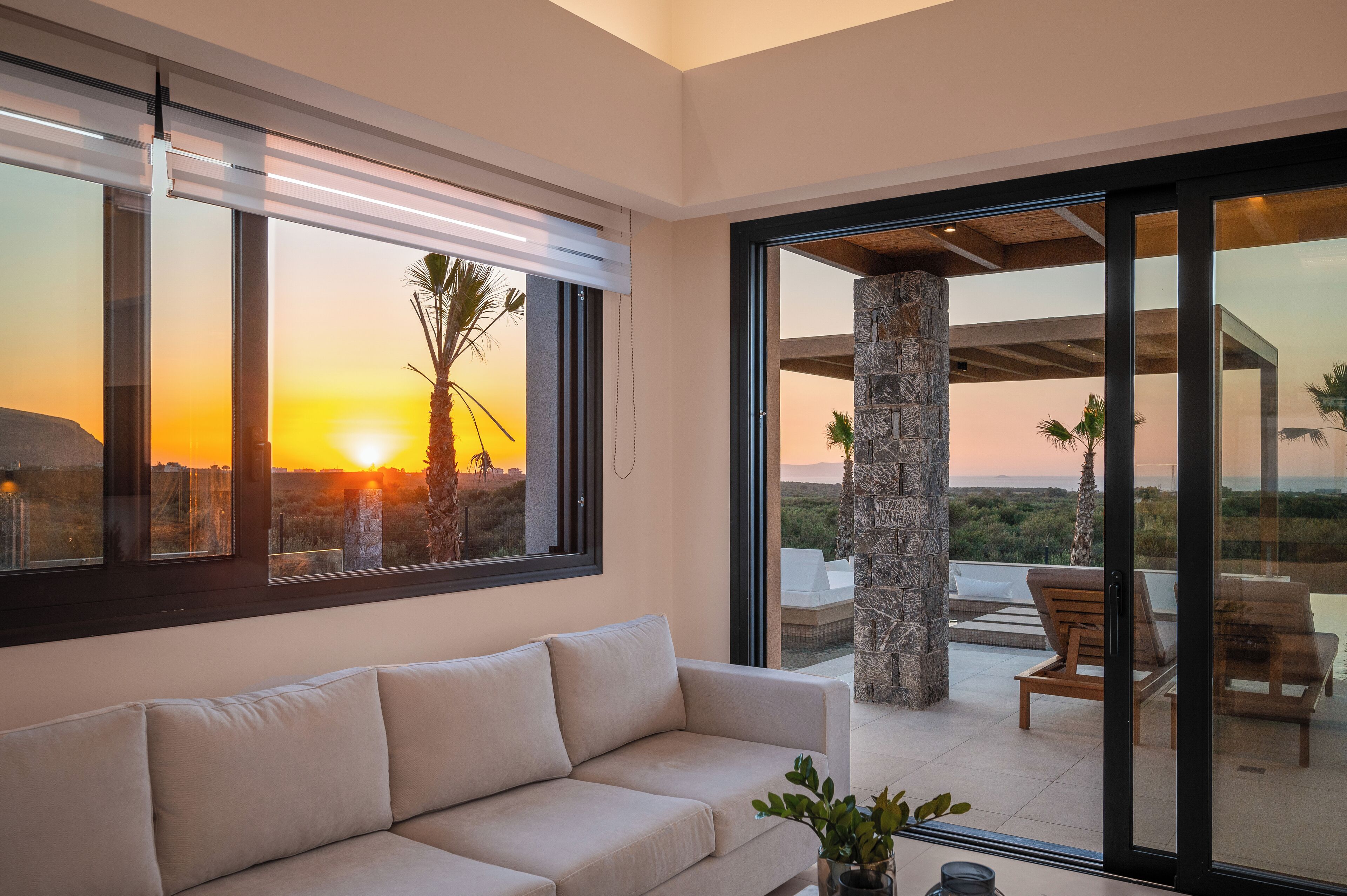 A modern living room with a sofa, large windows, and a view of a sunset and palm trees.