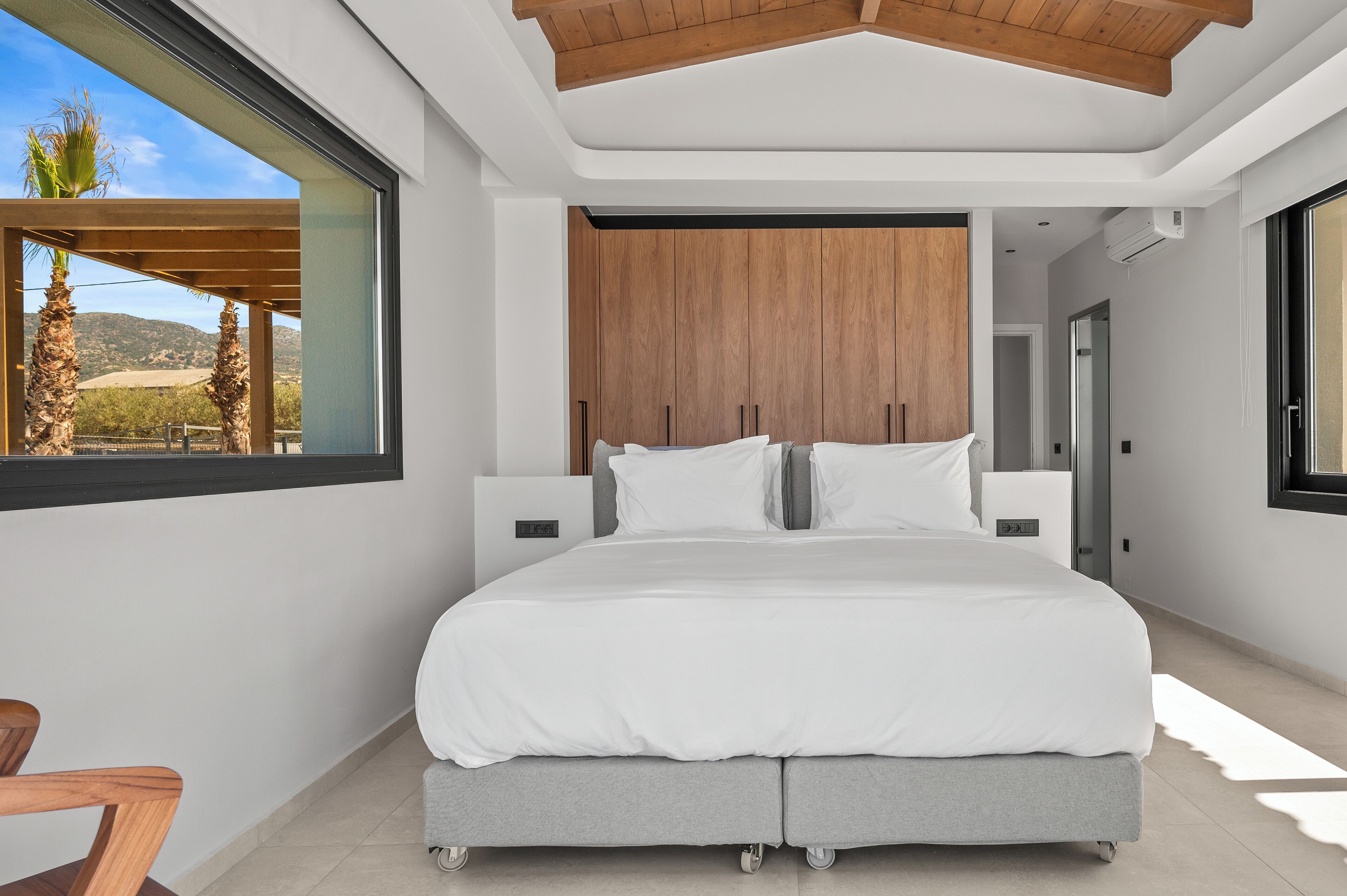 Ein modernes Schlafzimmer mit einem großen Bett, einem Holz-Kopfteil und Blick auf Palmen.