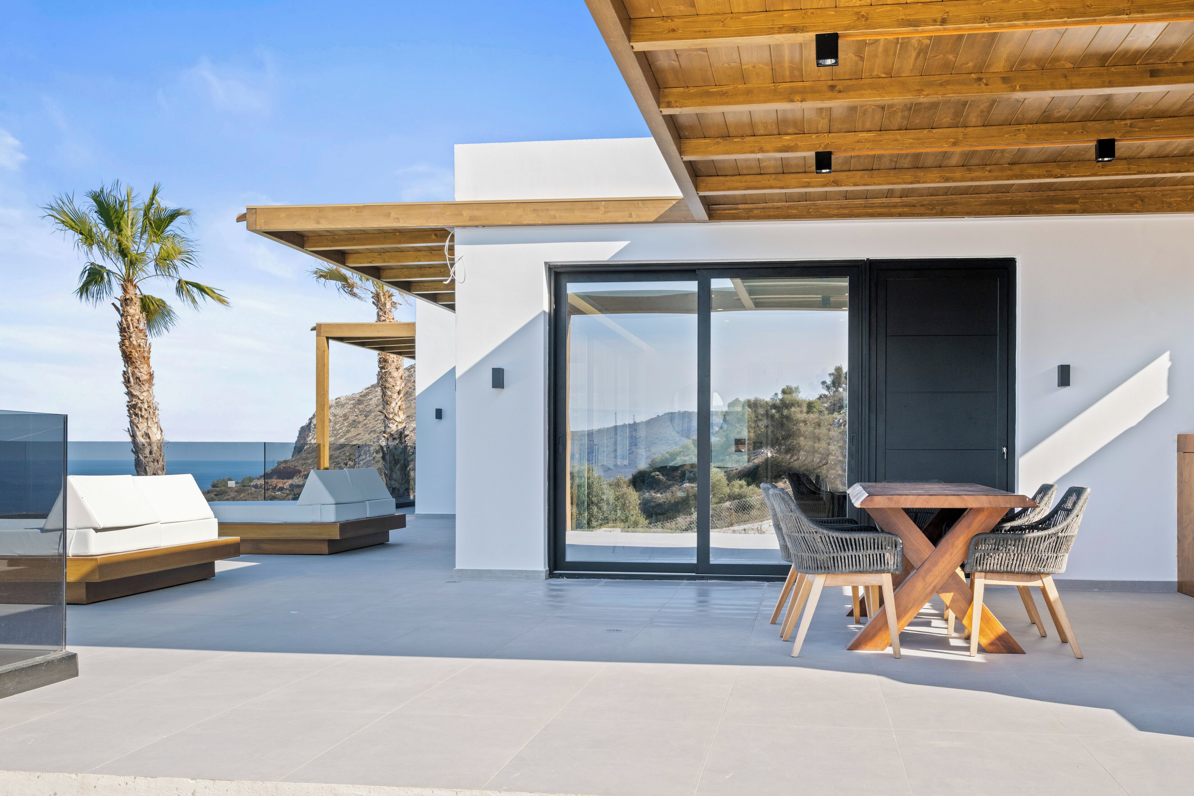A modern outdoor patio with a wooden ceiling, a glass door leading to a scenic view, and a dining area with wicker chairs and a wooden table.