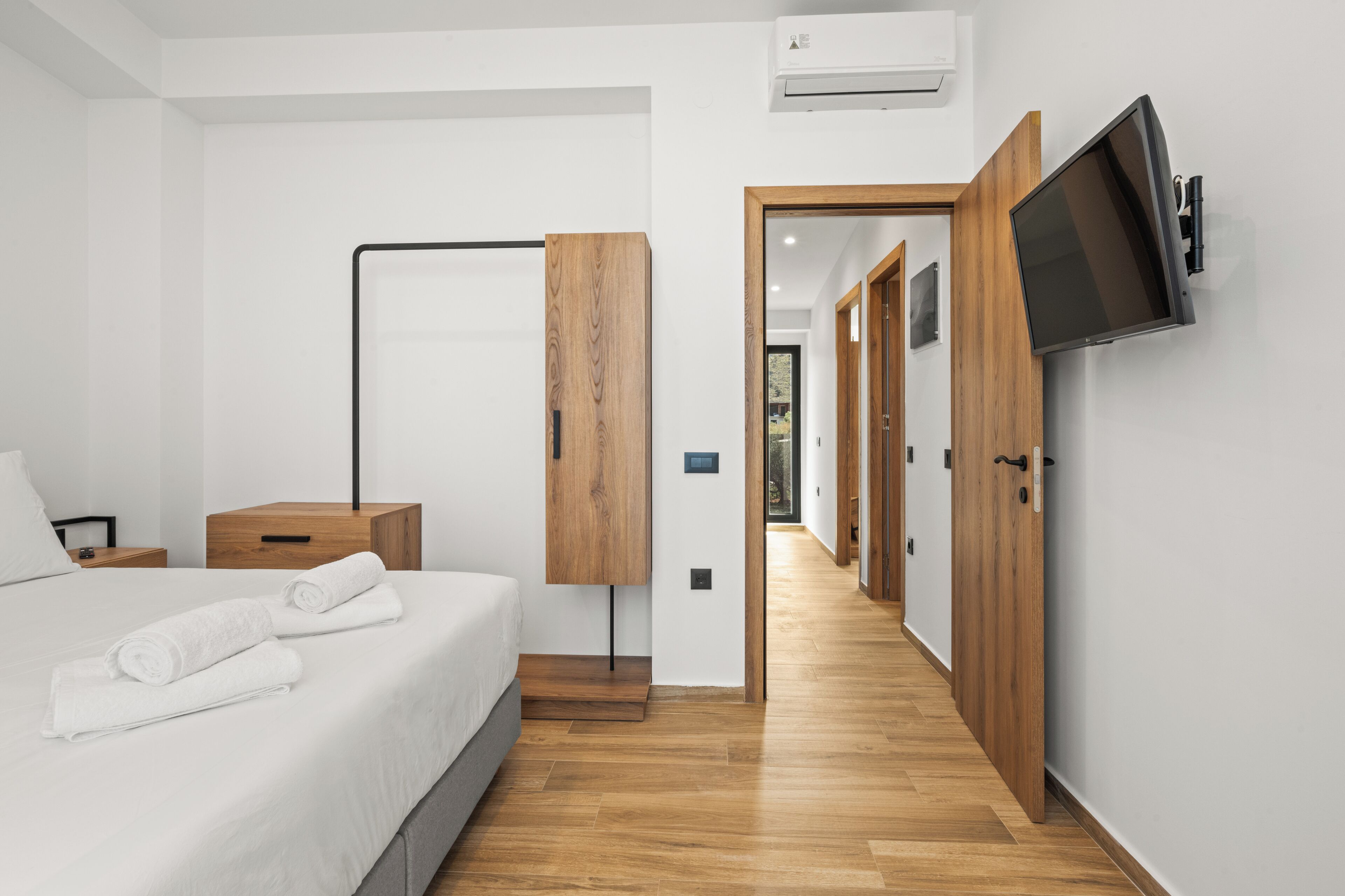 Une chambre d’hôtel moderne équipée d’un lit, d’un téléviseur à écran plat fixé au mur, d’un plancher en bois et d’un couloir menant à d’autres chambres.