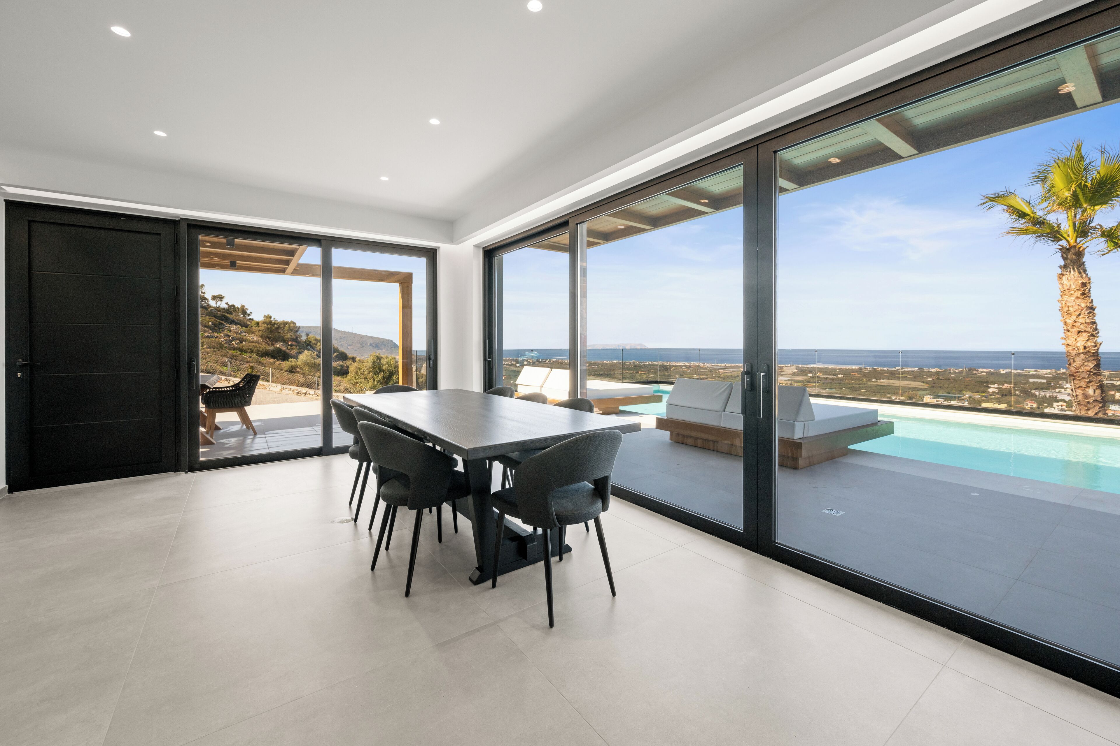 Un comedor moderno con una mesa grande y sillas, ventanas amplias con vista a una piscina y una palmera, y un panel corredizo que da a un patio exterior.