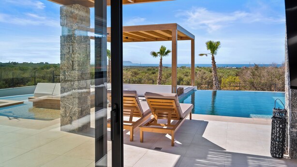 A modern outdoor pool area with lounge chairs, a stone pillar, and a view of the ocean. - Temma Villas (Hersonissos)