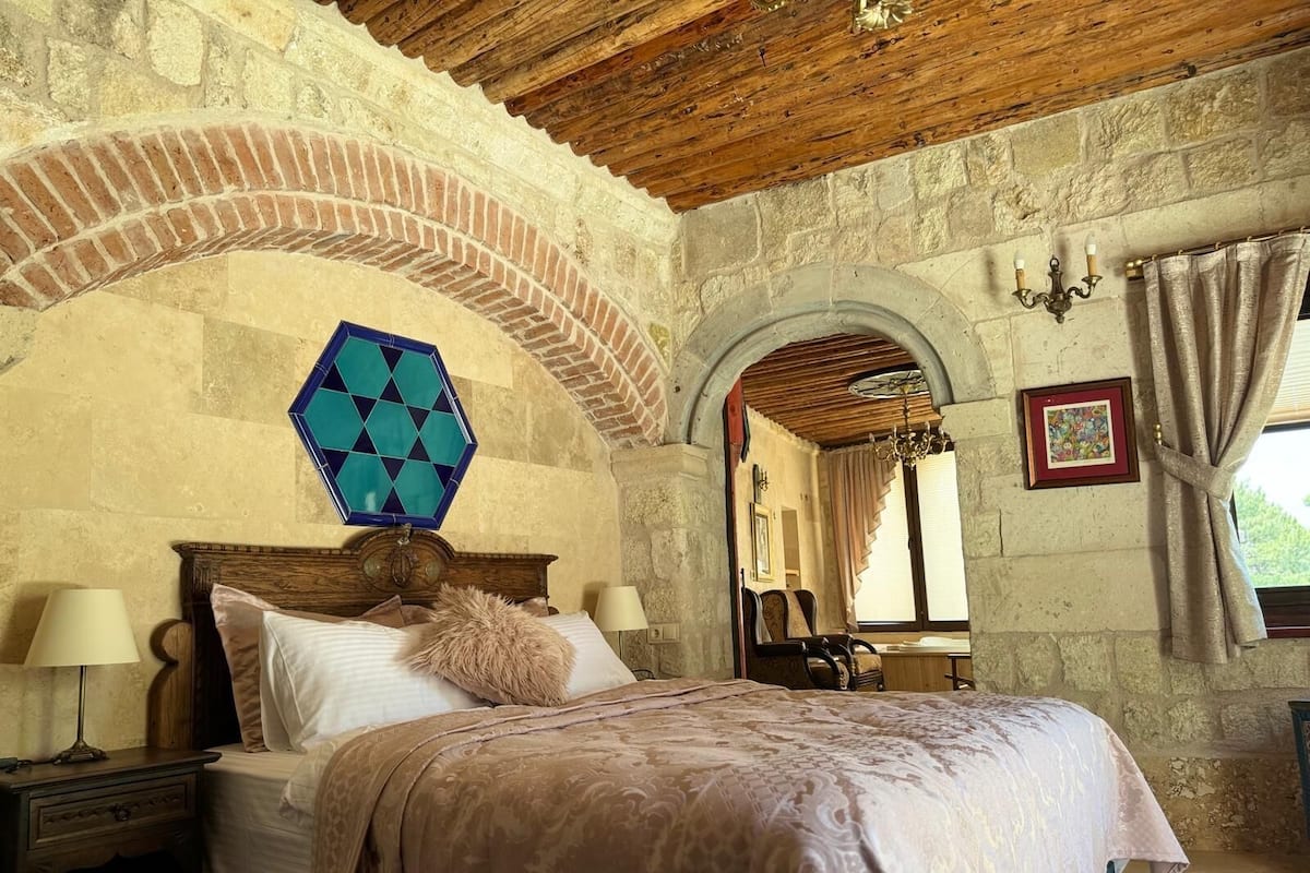 A bedroom with a stone wall, wooden ceiling, a bed with a patterned bedspread, a nightstand, a wall-mounted lamp, and a window with curtains.