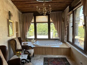 Quarto com teto de madeira, uma janela grande com cortinas, uma banheira, uma mesa de jantar com cadeiras e um lustre.