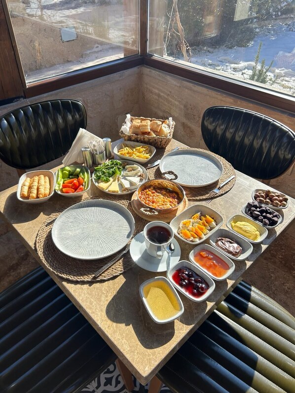 Uma mesa de pequeno-almoço com diversos pratos, incluindo frutas, queijos e pão, num quarto soalheiro com vista para uma paisagem coberta de neve.