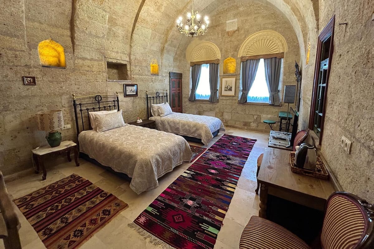 A room with two beds, a desk, and a chair. There are two windows with curtains, a chandelier, and a patterned rug on the floor.