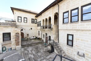 Pátio com edifícios de pedra, uma escadaria e uma pequena área externa com mesas e cadeiras.