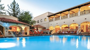A pool area with lounge chairs and umbrellas, a covered seating area with a railing, and a building with arched windows and a tiled roof.