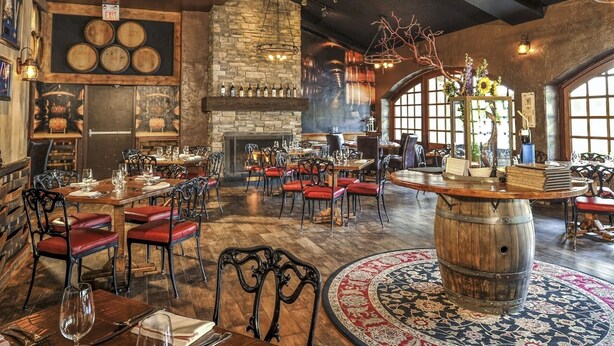 A restaurant with wooden tables, red cushioned chairs, a stone fireplace, and a large barrel centerpiece.