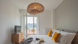 A bedroom with a bed, a dresser, a tray with breakfast, a hanging light, and a view of the city through a window. - Como Panorama in Como (Como)