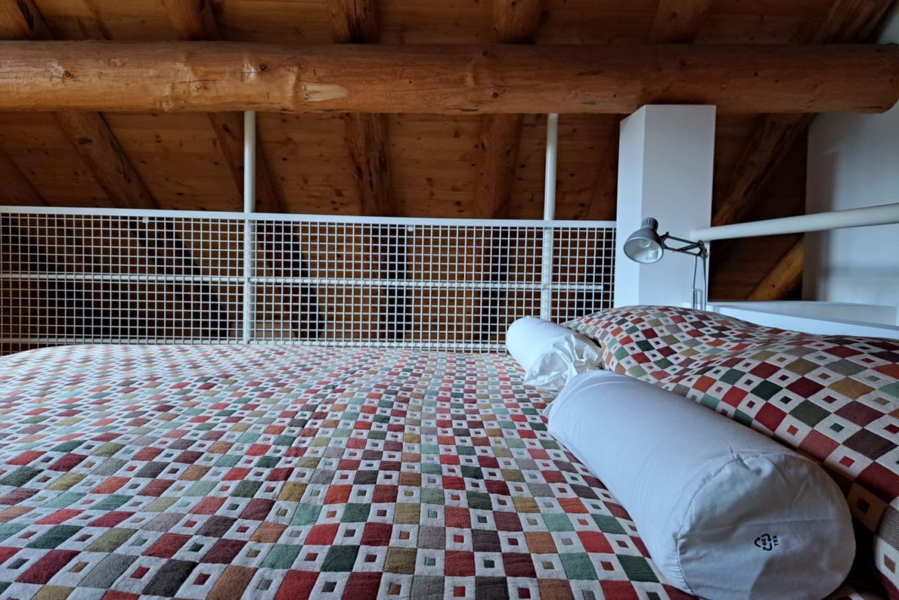 Habitación con techo de madera, colcha estampada, almohada blanca y pared blanca con ventana.