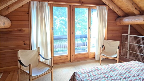 Une pièce de cabane en bois comprenant un lit, deux chaises et une grande fenêtre avec des rideaux.