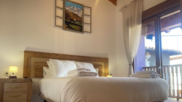 A bedroom with a wooden headboard, a nightstand with a lamp, a bed with white bedding, and a painting of mountains on the wall. - Charming rural house Casa Zalo for 2-4 people in Mestas de Con (Cangas de Onís)