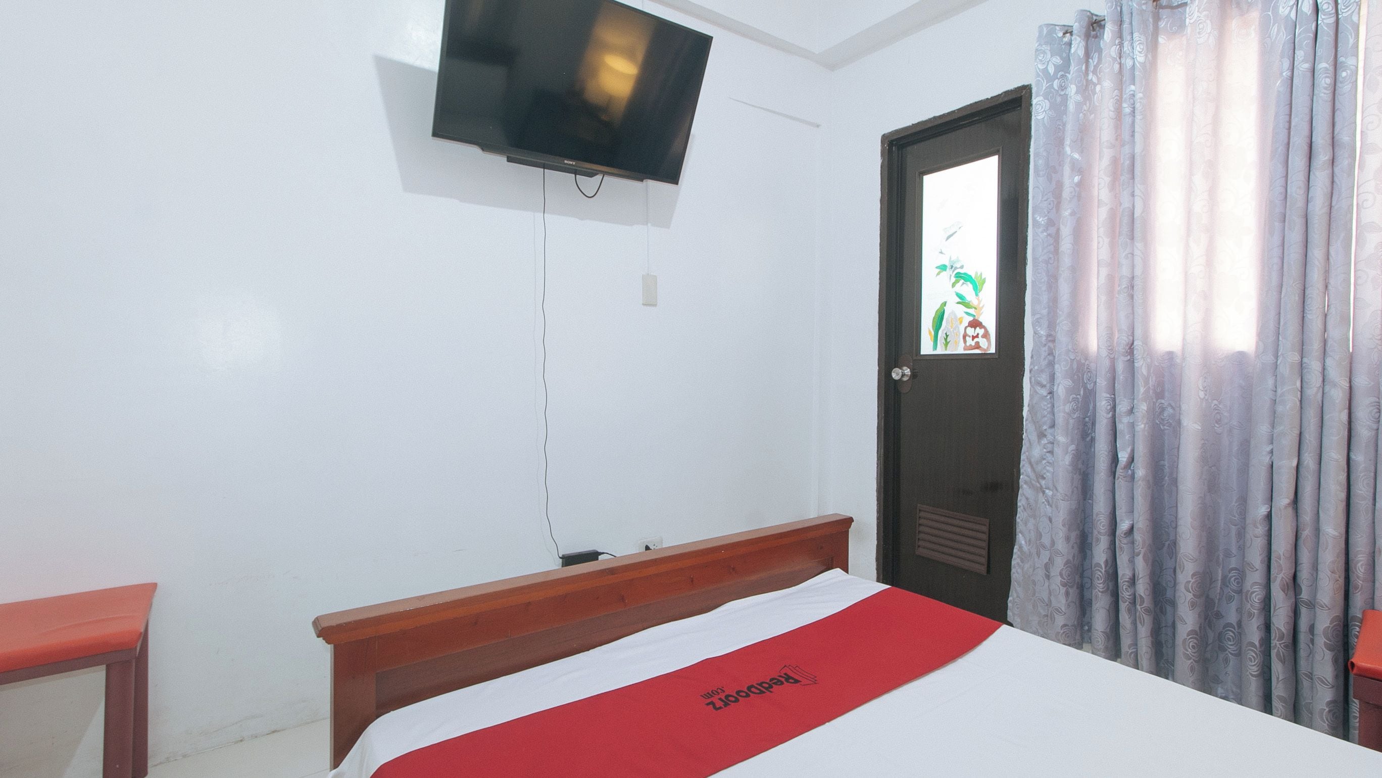 A hotel room with a bed, a television mounted on the wall, a door with a window, and a curtain.