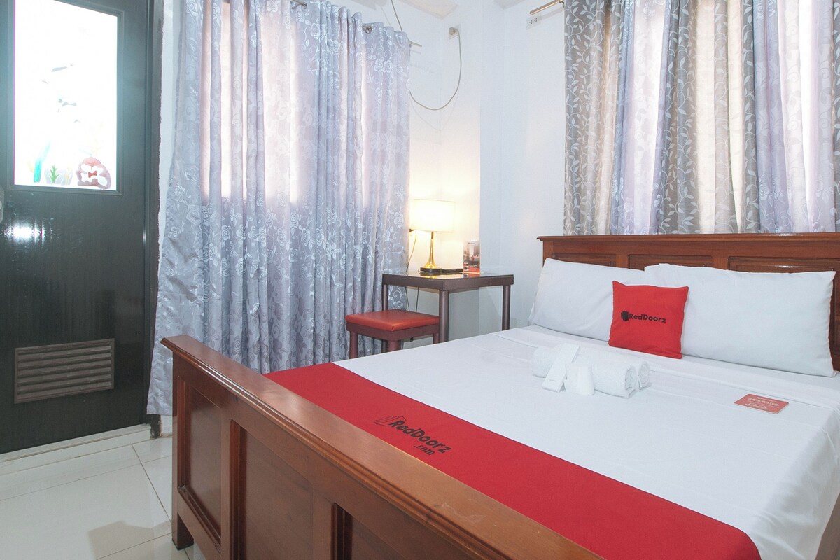Habitación de hotel con cama de madera, colcha roja, almohada roja, mesita de noche con lámpara y cortinas con estampado floral.