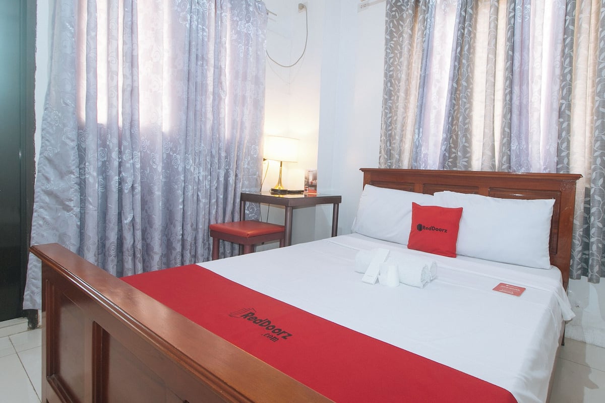 Une chambre d’hôtel avec un lit en bois, une couverture rouge et un oreiller orné d’un logo rouge.