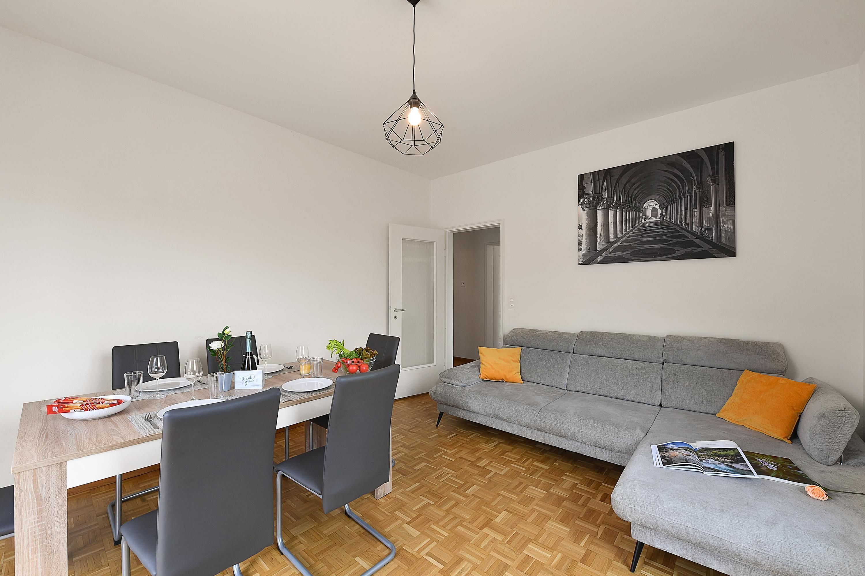 Un coin repas avec une table dressée pour quatre personnes, un canapé gris agrémenté de coussins orange et une œuvre d’art murale représentant un couloir.
