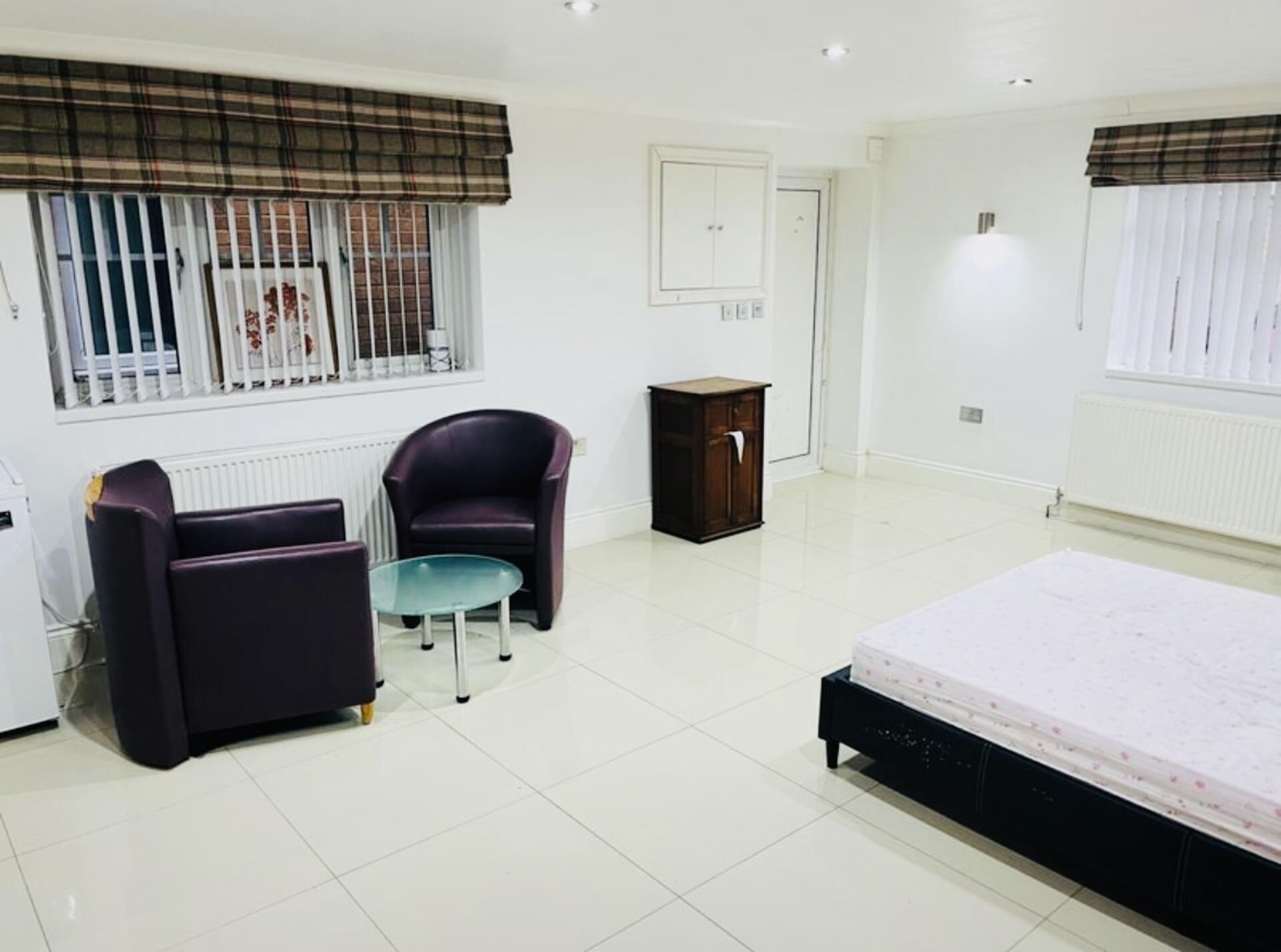 A bedroom with a bed, two armchairs, a small table, a cabinet, and a window with blinds.