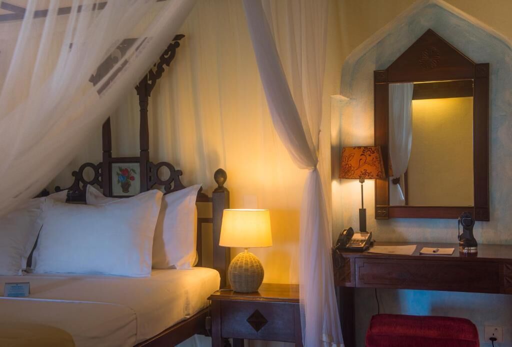 A hotel room with a bed, bedside table with a lamp, a desk with a telephone, and a mirror.