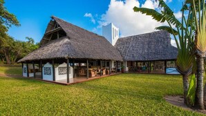 A thatched-roof building with an attached white structure, surrounded by a well-maintained lawn and palm trees. - Amani Beach Resort  (Kutani)