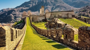 A historic stone wall with a grassy pathway leading to a fortified structure with towers, set against a mountainous backdrop. - Bellinzona in Centro (Bellinzona)