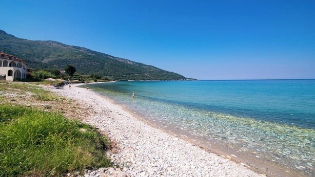 A pebble beach with clear water, a building with arches, and a mountain in the background. - Kapsogeorgis Rooms (Thasos)