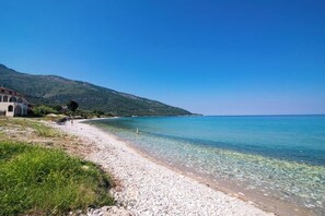 A pebble beach with clear water, a building with arches, and a mountain in the background. - Kapsogeorgis Rooms (Thasos)