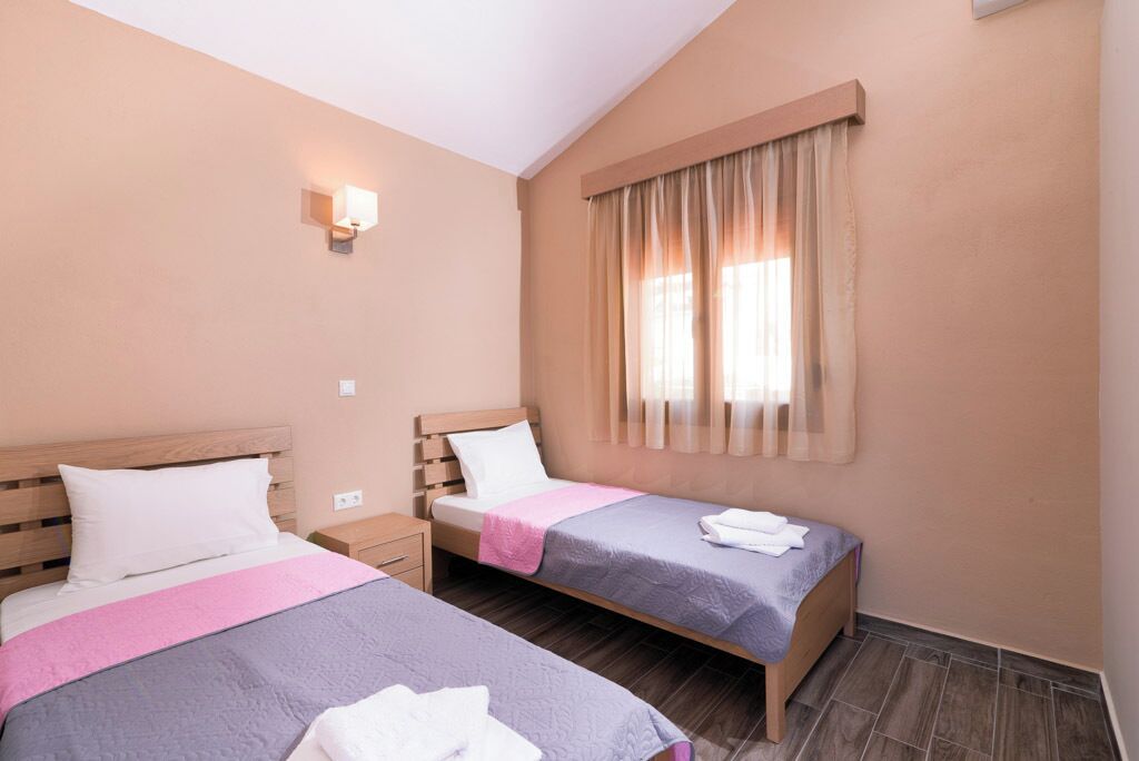 Habitación de hotel con dos camas, una mesita de noche, un ventanal con cortinas e iluminación empotrada en la pared.