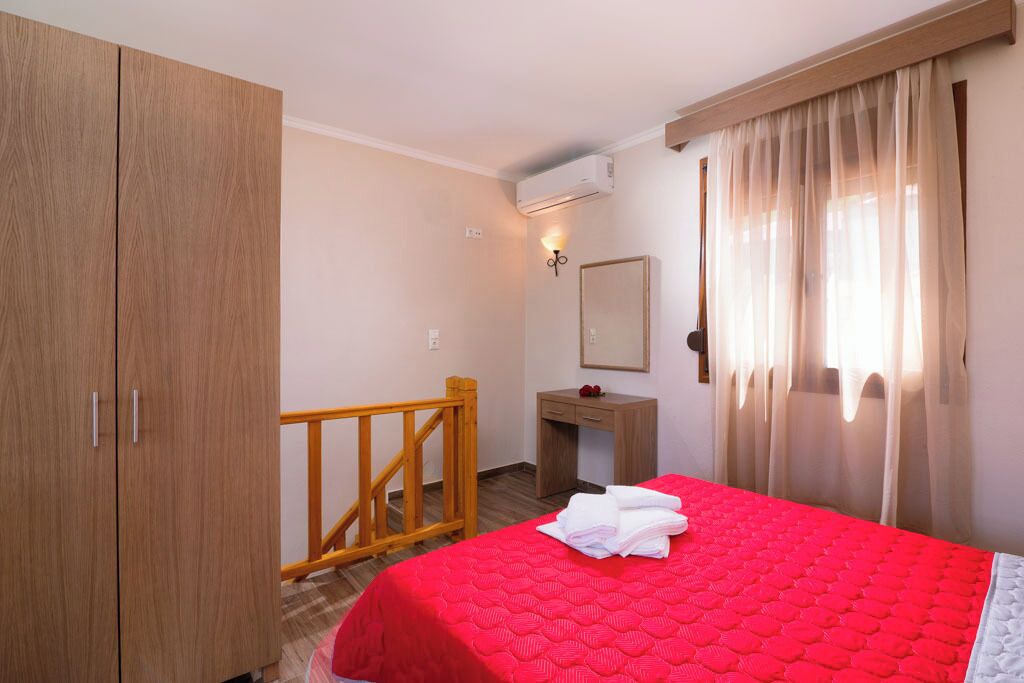 A hotel room with a wooden wardrobe, a desk with a mirror, a bed with a red quilt, and a window with sheer curtains.