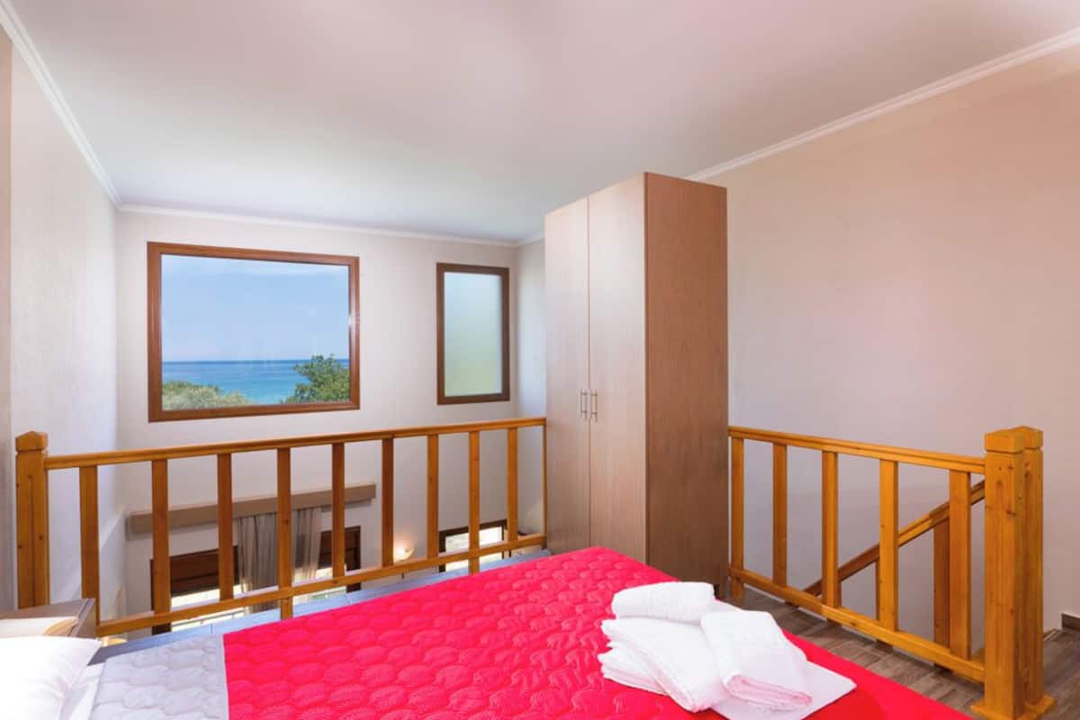 A bedroom with a bed, a window with a view of the sea, a wooden wardrobe, and a balcony railing.