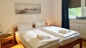 Ein Doppelbett mit weißen Bettwäsche, ein Nachttisch mit Lampe, ein Gemälde einer Strandansicht und ein Fenster mit Vorhängen.