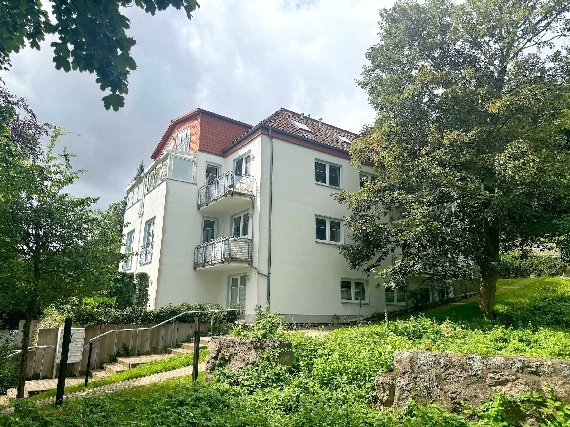 Un edificio residencial de varios pisos con balcones, rodeado de abundante vegetación y un muro de piedra.