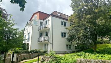 Un edificio residencial de varios pisos con balcones, rodeado de abundante vegetación y un muro de piedra.