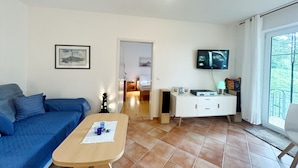 Ein Wohnzimmer mit einem blauen Sofa, einem Holztisch, einem an der Wand befestigten Fernseher und einem großen Fenster.
