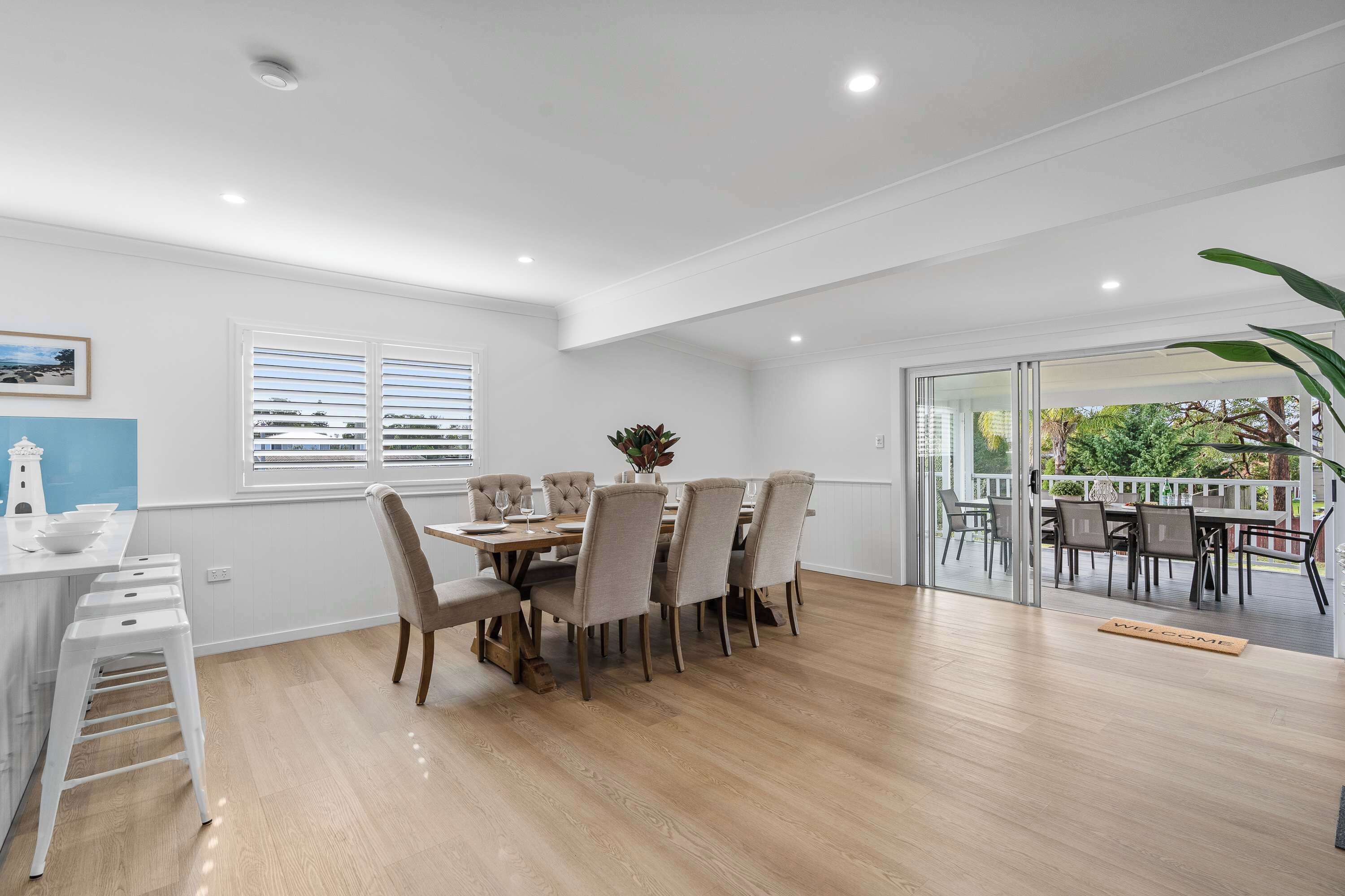Un comedor con mesa y sillas de madera, una pared blanca con un cuadro, y vista a un patio exterior por una puerta de vidrio.