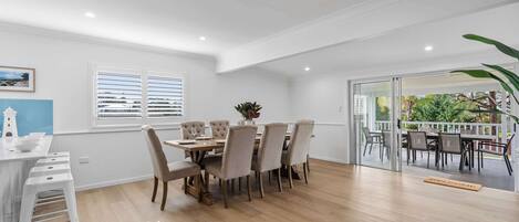 Un comedor con mesa y sillas de madera, una pared blanca con un cuadro, y vista a un patio exterior por una puerta de vidrio.
